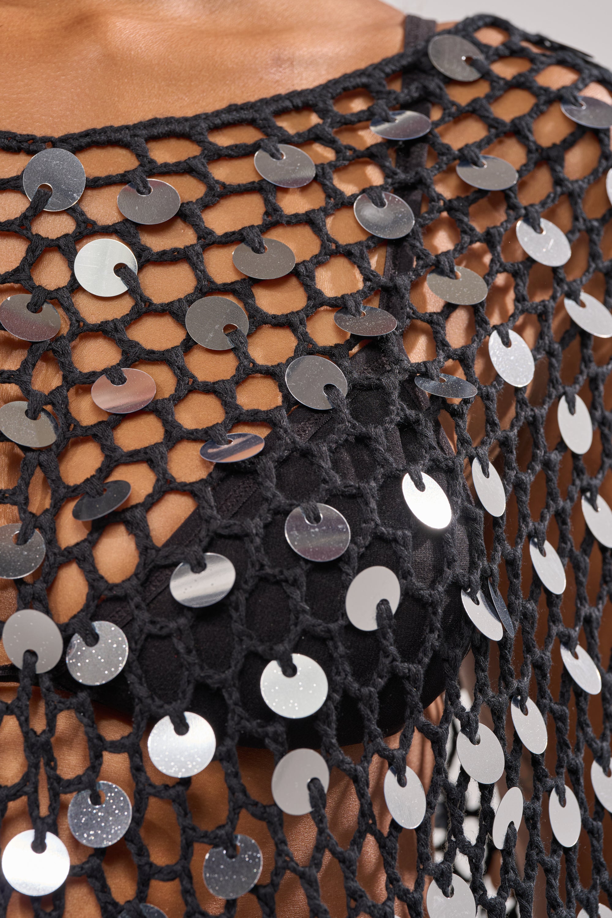 Close-up of a person wearing the UNDER THE LIGHTS CROCHET SEQUIN PONCHO, featuring large, shiny silver sequins and intricate black netted crochet over a black bra. The image highlights the poncho's textured sparkle and eye-catching design. 6