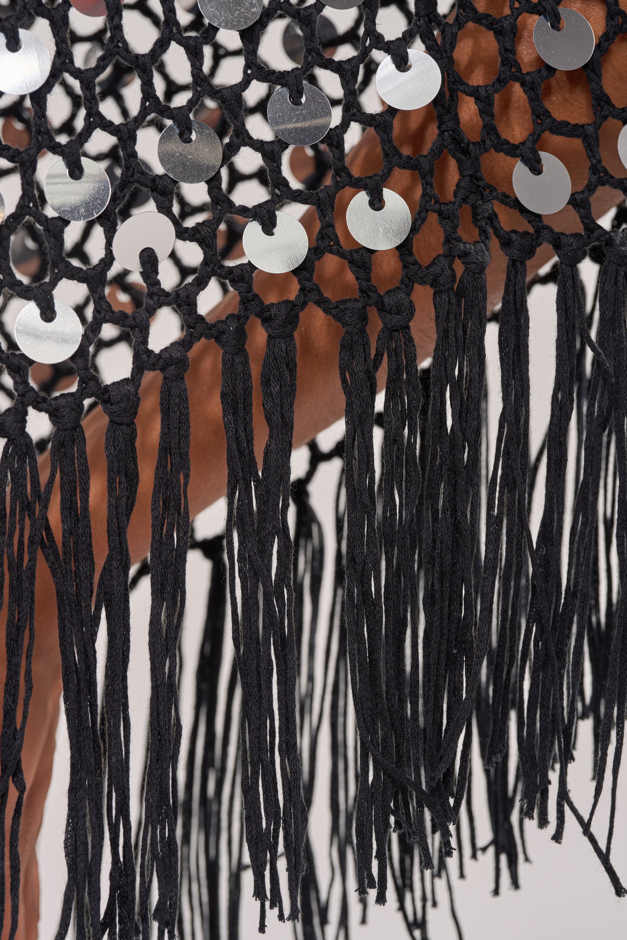 Close-up of the UNDER THE LIGHTS CROCHET SEQUIN PONCHO, showcasing black crochet with shiny silver sequins and long fringe trim, elegantly draped over a brown surface. 7