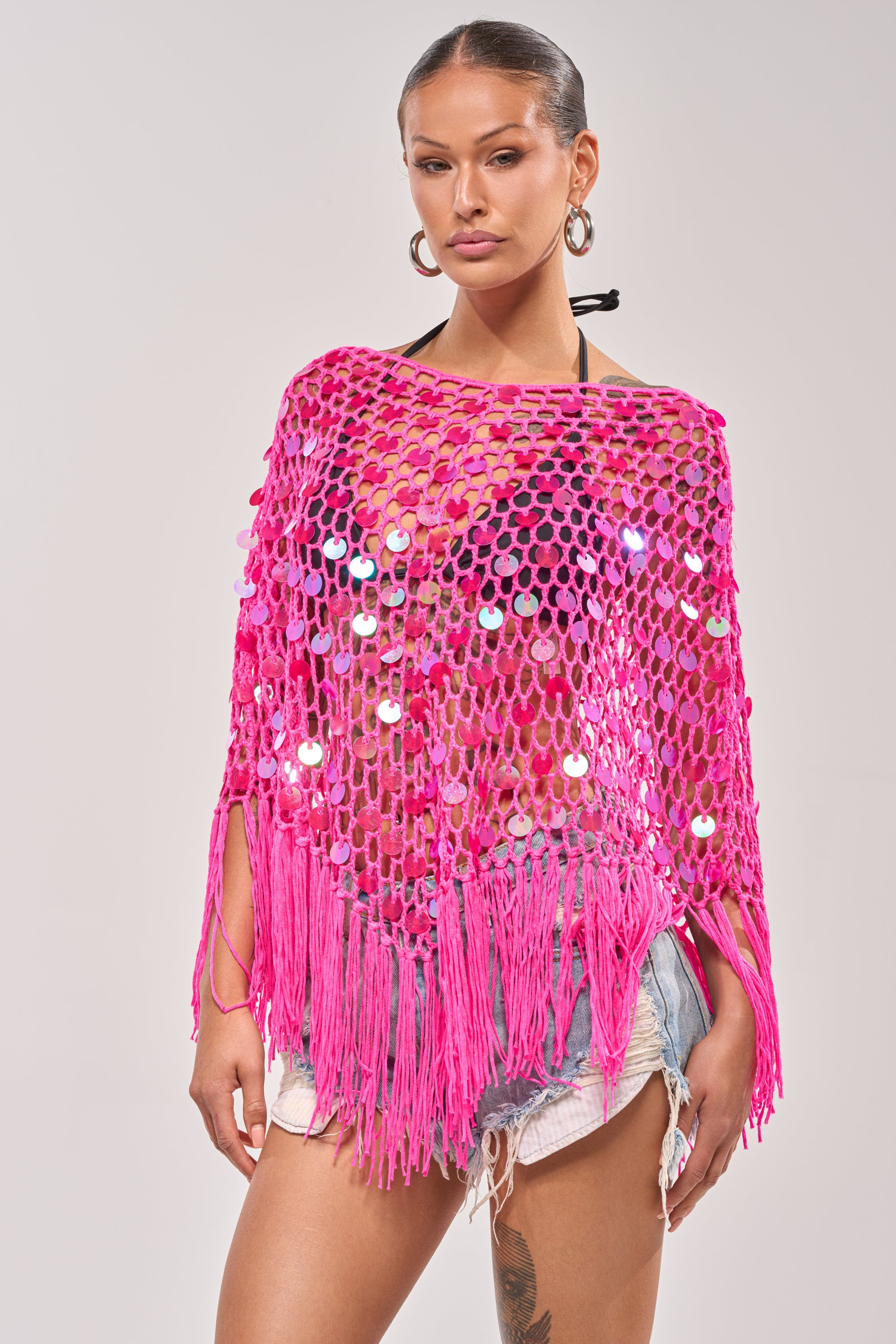 A woman wears the ITS MY WORLD CROCHET SEQUIN PONCHO in hot pink over a bikini top and denim shorts, standing against a plain light background. 0