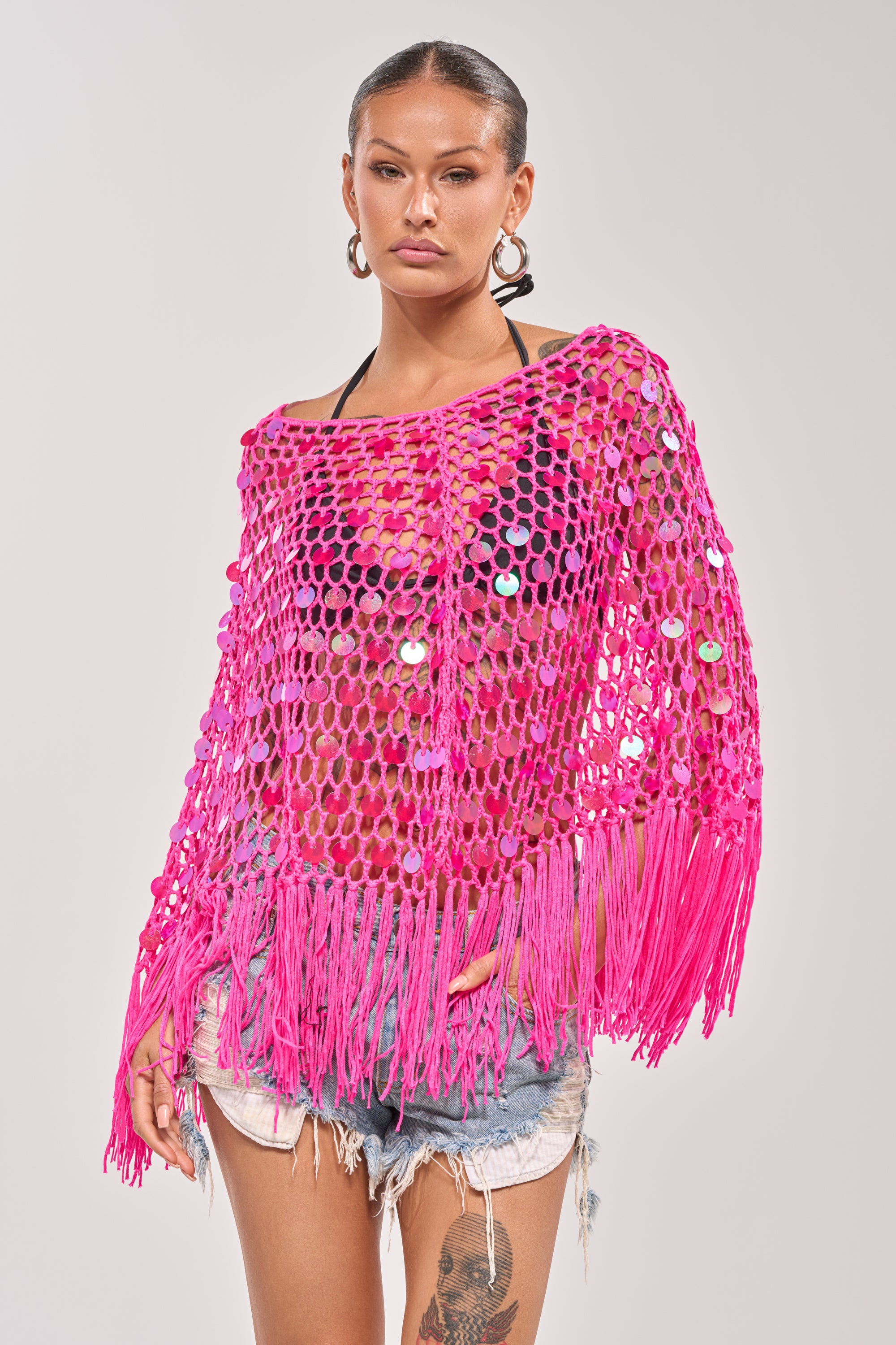 A woman models the ITS MY WORLD CROCHET SEQUIN PONCHO—hot pink with fringe and shimmering sequins—layered over a black bikini top and distressed denim shorts, standing against a plain light background. 1