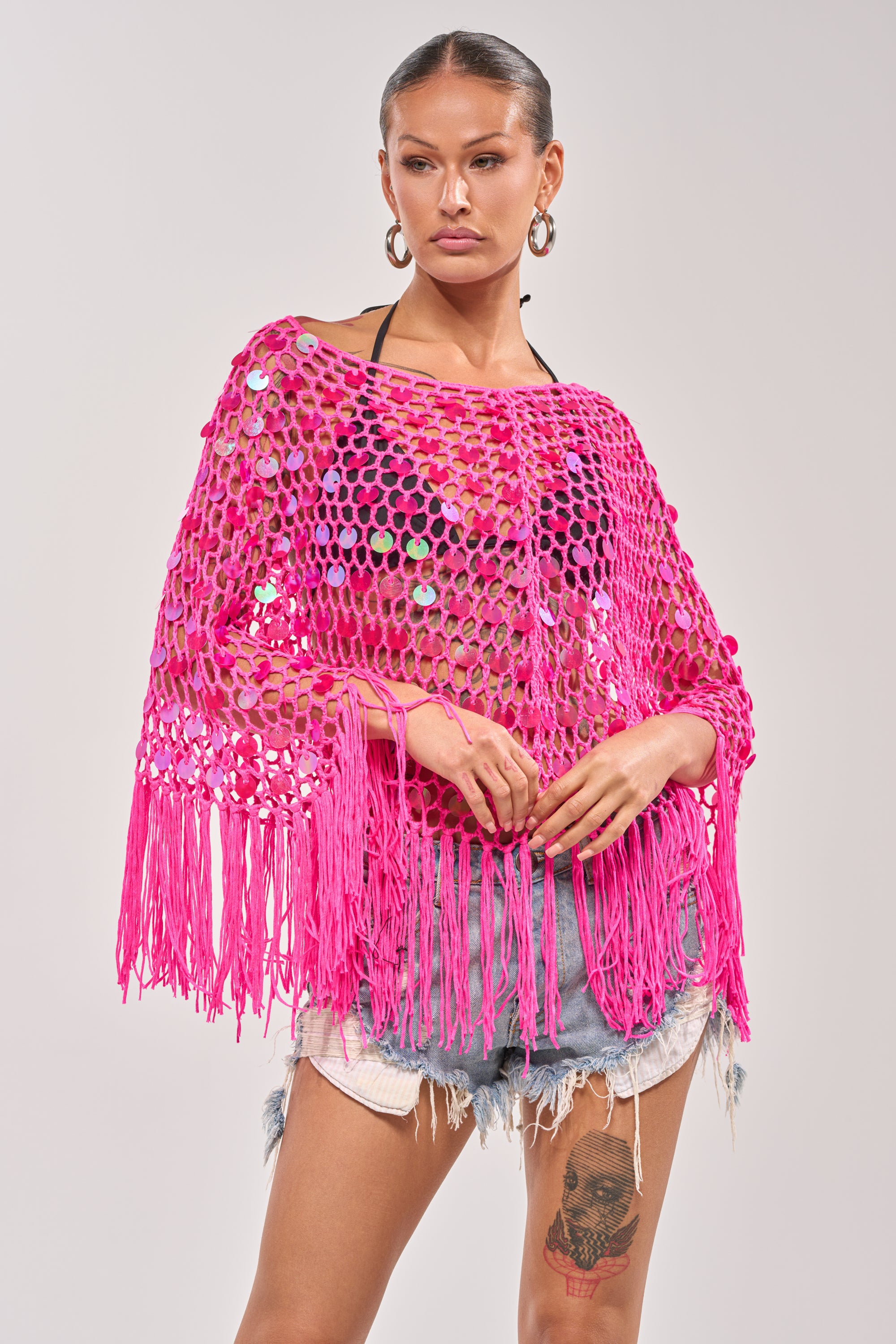 Model wearing the ITS MY WORLD CROCHET SEQUIN PONCHO—bright pink with sequins and fringe—styled over a black bikini top and distressed denim shorts, posing against a plain background. 2
