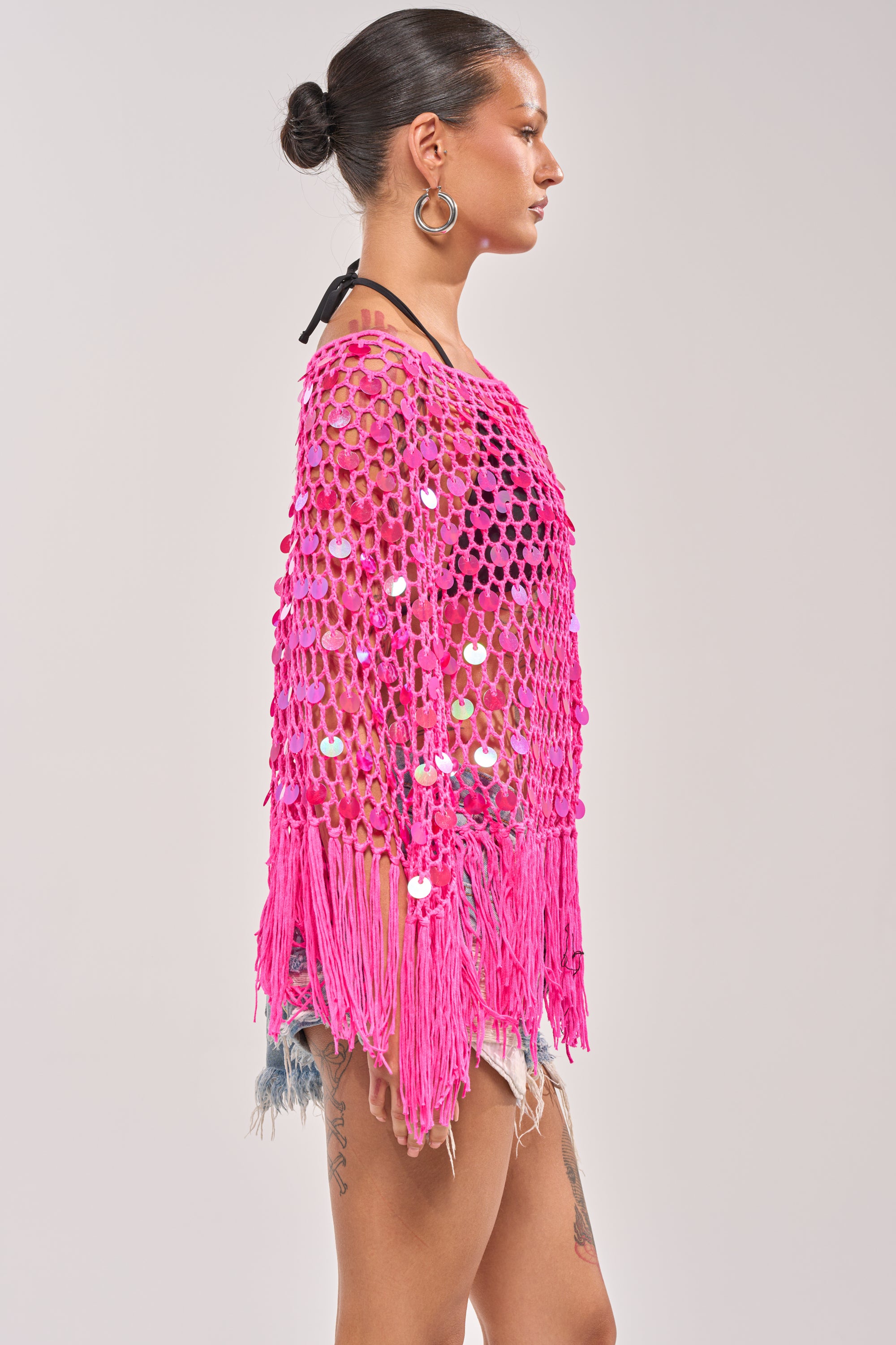 A person with hair in a bun models the ITS MY WORLD CROCHET SEQUIN PONCHO—vibrant pink with sequins and fringe—over a black top and denim shorts, posing in profile against a plain background. 3