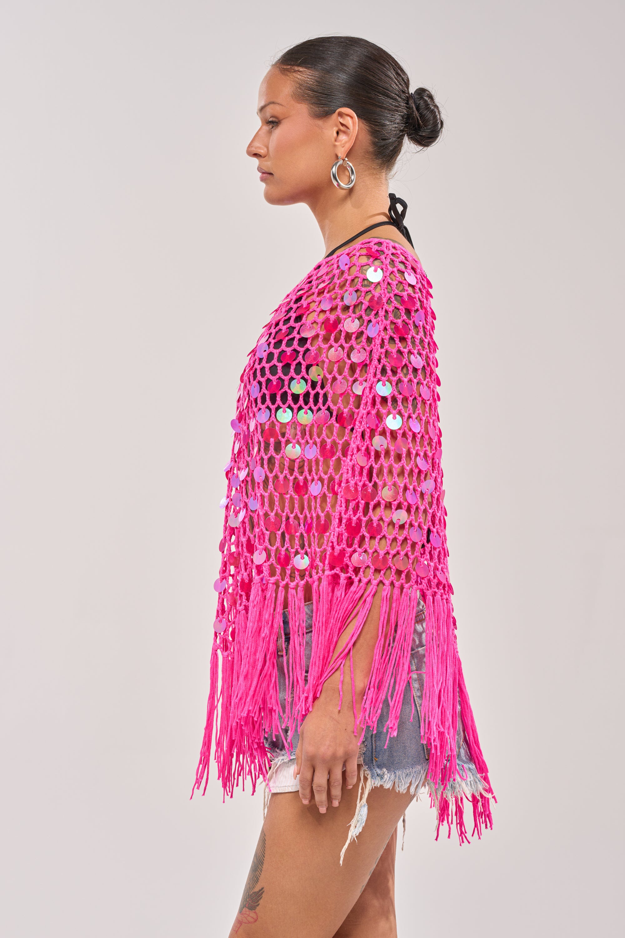 A woman in profile wears the ITS MY WORLD CROCHET SEQUIN PONCHO, a vibrant pink festival piece with shiny sequins, styled over a black top and denim shorts. She pulls her dark hair back and accessorizes with hoop earrings. 4