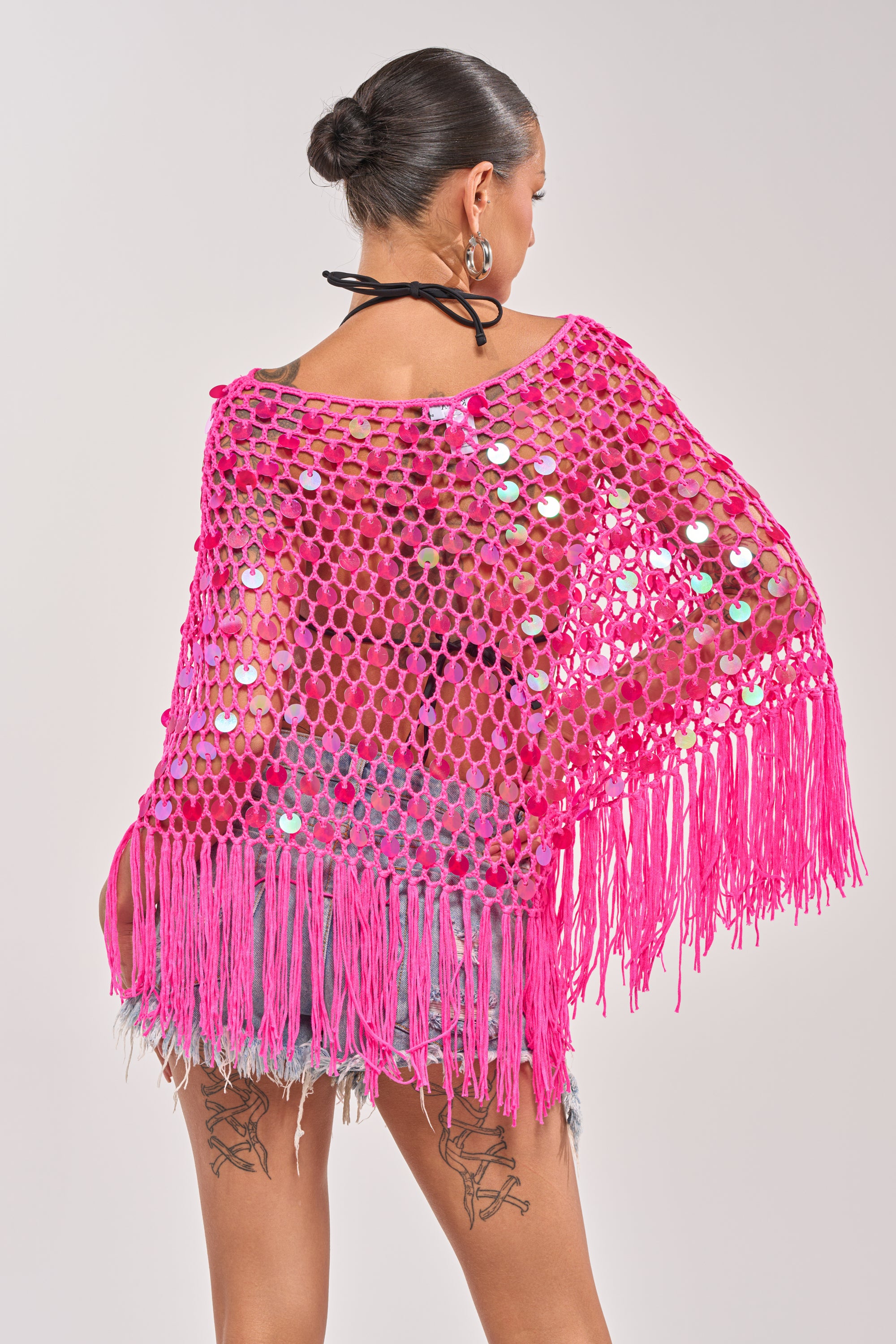 A woman with thigh tattoos wears the ITS MY WORLD CROCHET SEQUIN PONCHO in bright pink over denim shorts. Facing away, her hair in a neat bun and a black halter strap show off this festival-ready style’s unique flair. 5