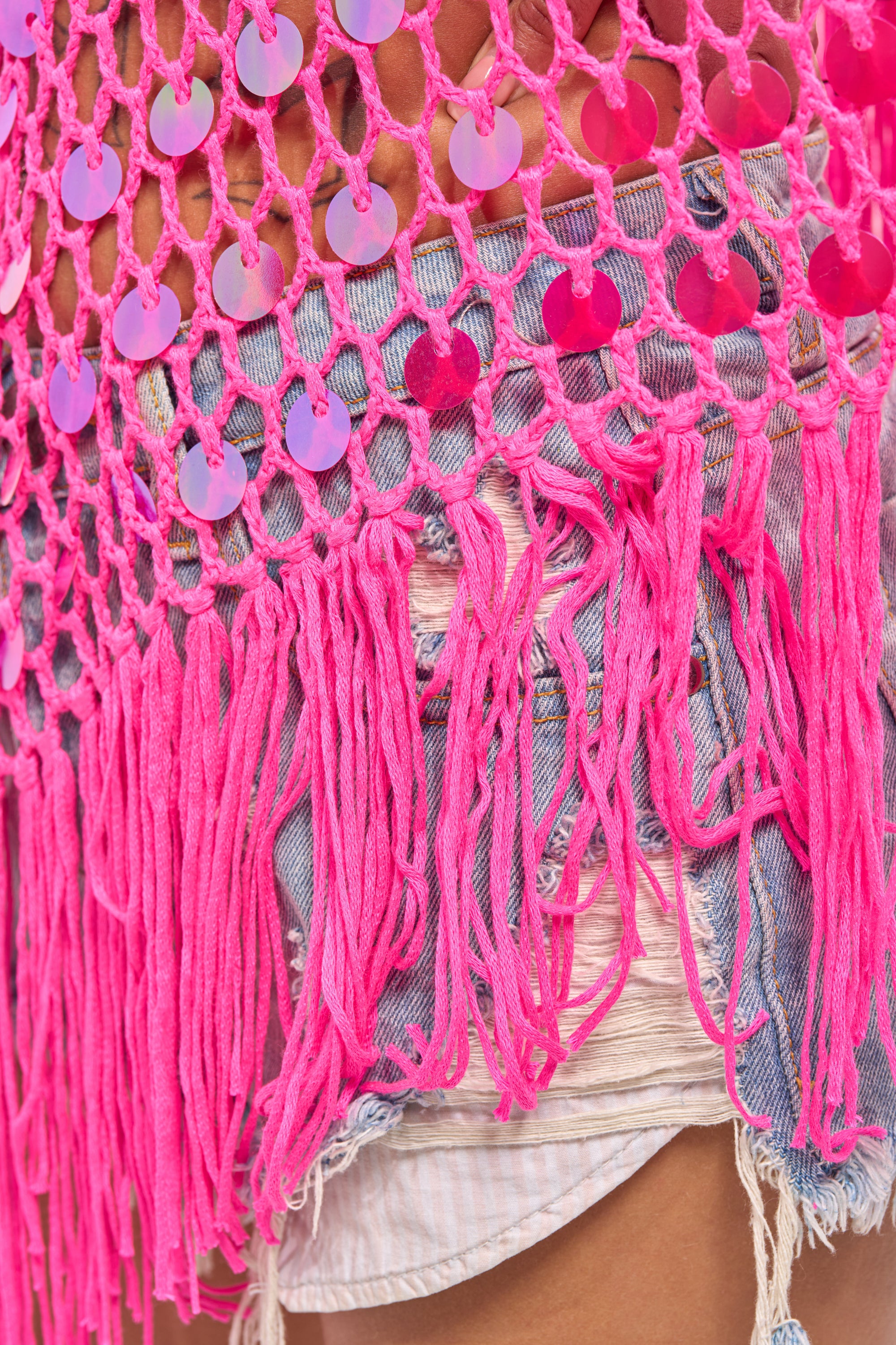 Close-up of a person wearing frayed denim shorts and the ITS MY WORLD CROCHET SEQUIN PONCHO, featuring pink crochet, fringe, and shiny pink sequins—perfect for festival style. 8