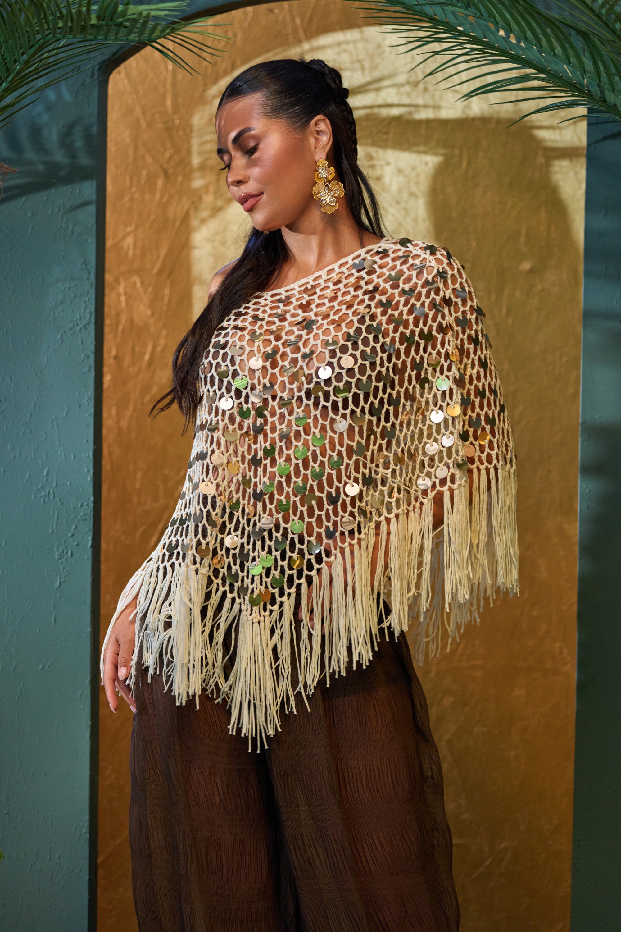 A woman poses before a gold and teal backdrop, wearing the BELLA CROCHET SEQUIN PONCHO with fringe, large gold earrings, and sheer brown pants. Palm leaves frame the top of the scene.