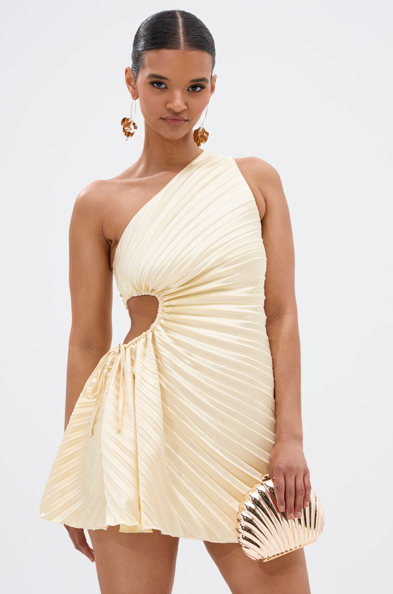 A woman wears the MAKE IT WORK PLEATED MINI DRESS IN YELLOW, featuring an asymmetrical one-shoulder design, pleats, and a side cutout. She holds a metallic shell clutch, with slicked-back hair and bold gold statement earrings.