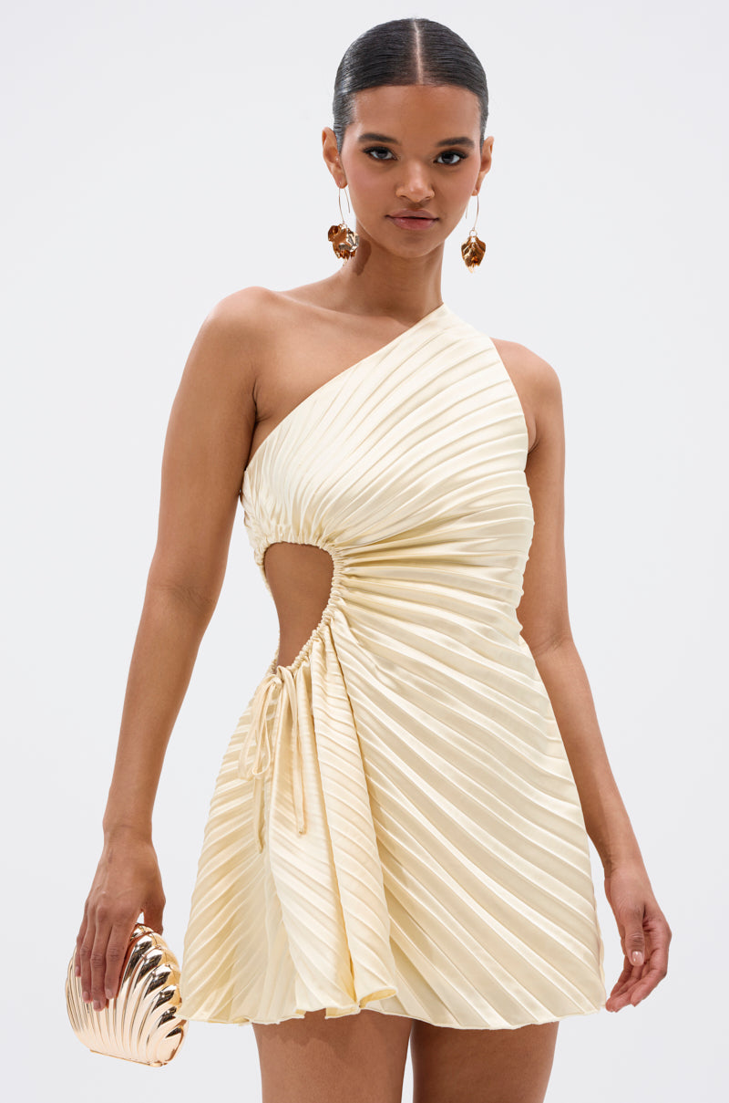 A woman wears the MAKE IT WORK PLEATED MINI DRESS IN YELLOW, featuring an asymmetrical one-shoulder design, pleats, side cutout, and drawstring detail. She accessorizes with a gold clutch and statement earrings, hair pulled back for a chic touch.