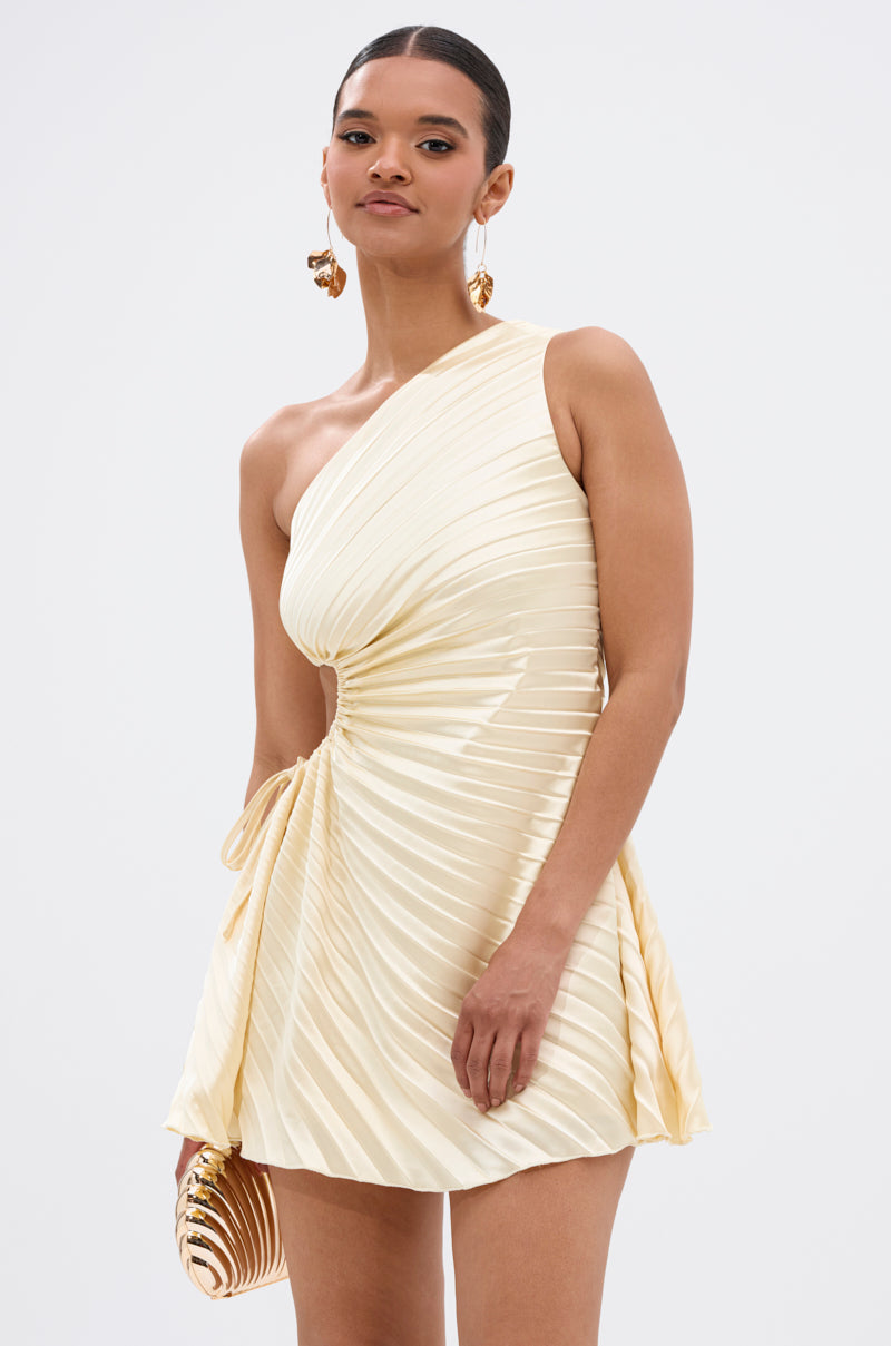 A woman models the MAKE IT WORK PLEATED MINI DRESS IN YELLOW—an asymmetrical one-shoulder, yellow pleated mini dress with ruched side detail—styled with a gold striped clutch and bold statement earrings against a plain backdrop.