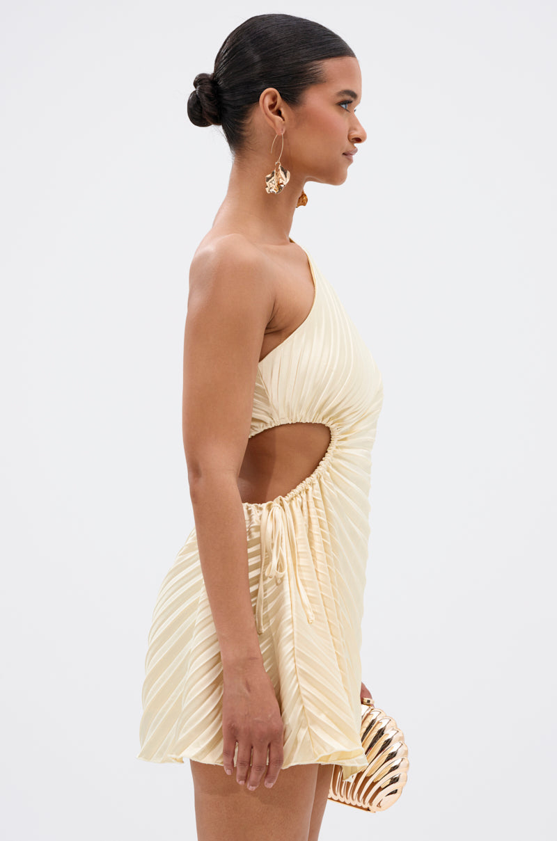 A woman stands in profile against a plain background wearing the MAKE IT WORK PLEATED MINI DRESS IN YELLOW, featuring an asymmetrical one-shoulder design, pleats, and a side cut-out. She accessorizes with gold earrings, a gold clutch, and a sleek bun.