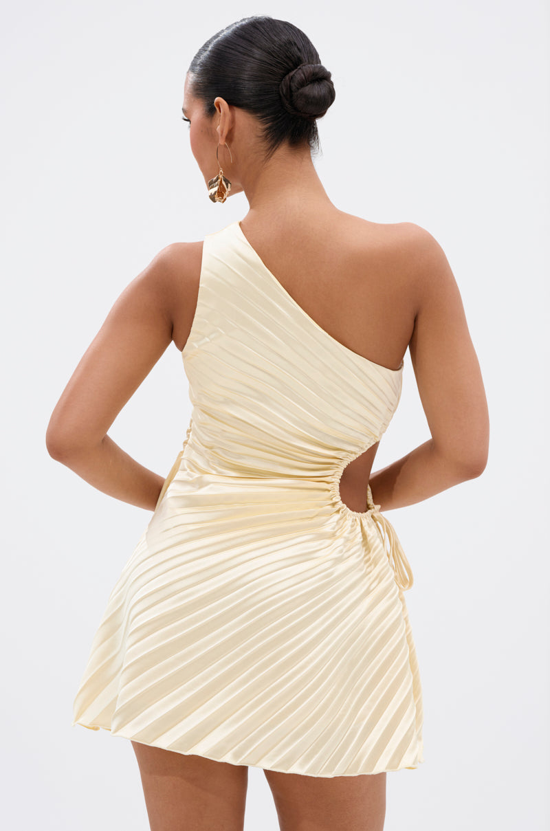 A woman with dark hair in a sleek bun models the MAKE IT WORK PLEATED MINI DRESS IN YELLOW, showing off its shiny pleats, asymmetrical one-shoulder design, and side cutout as she stands with her back to the camera against a light background.