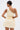 A woman with dark hair in a sleek bun models the MAKE IT WORK PLEATED MINI DRESS IN YELLOW, showing off its shiny pleats, asymmetrical one-shoulder design, and side cutout as she stands with her back to the camera against a light background.