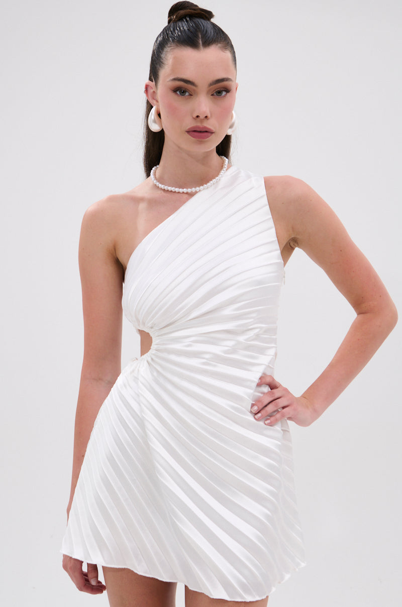 A woman models the MAKE IT WORK PLEATED MINI DRESS IN WHITE, styled with a pearl necklace and large white hoop earrings, standing against a plain background with one hand on her hip. 0