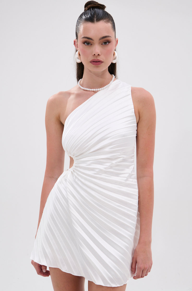 A woman stands against a plain white background wearing the MAKE IT WORK PLEATED MINI DRESS IN WHITE—an asymmetrical one-shoulder mini dress with waist cutout and pleats. She has long, straight hair, large hoop earrings, and a pearl necklace. 1