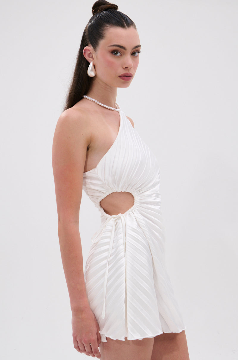 A woman poses in the MAKE IT WORK PLEATED MINI DRESS IN WHITE, featuring a pleated mini silhouette, asymmetrical one-shoulder design, and circular side cut-out. She accessorizes with pearl jewelry against a plain white background. 2