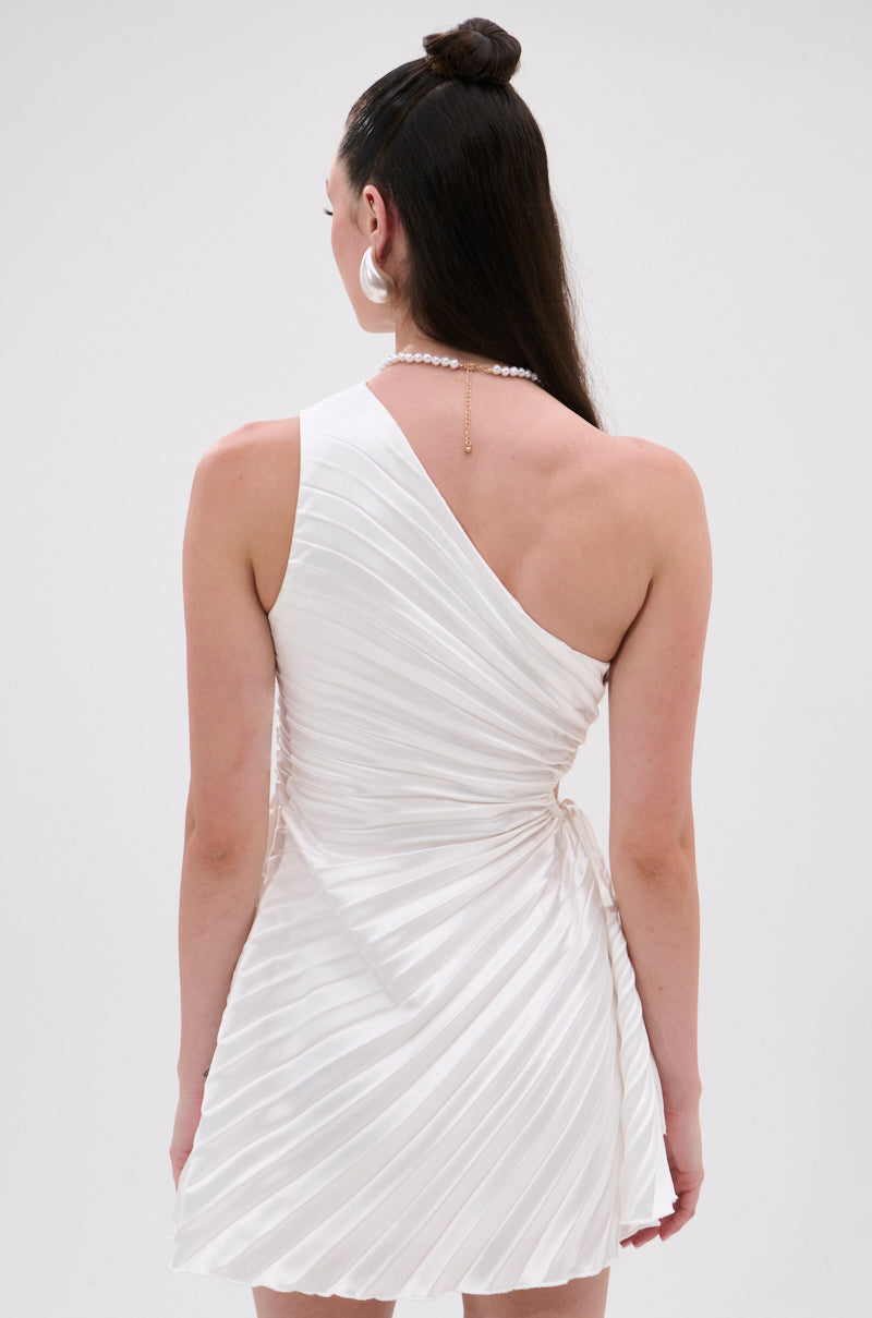 A woman with long dark hair styled half up wears the Make It Work Pleated Mini Dress in White, featuring an asymmetrical one-shoulder design and pleats. She accessorizes with large hoop earrings and a drop-chain necklace draped down her back. 4