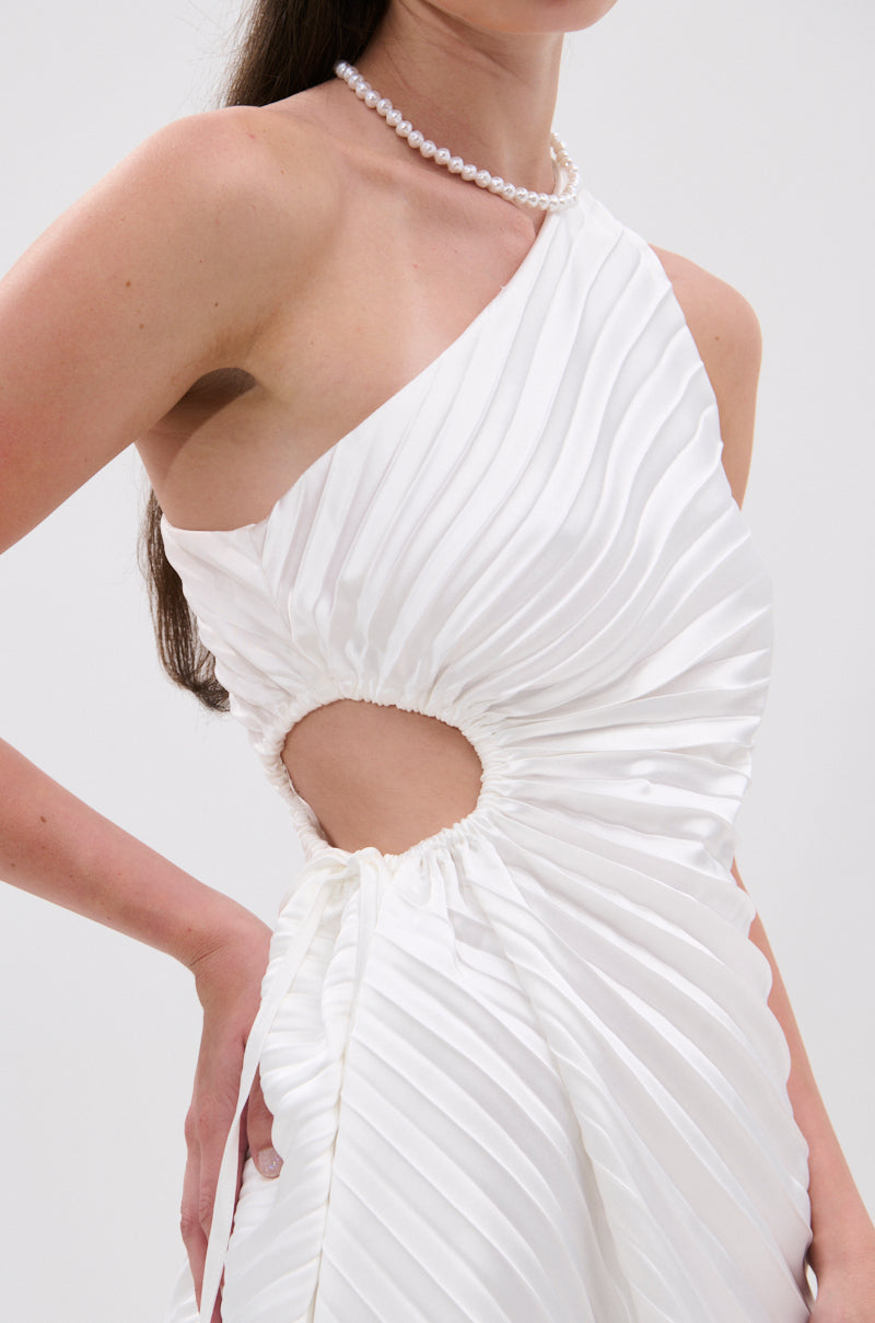 A person wears the MAKE IT WORK PLEATED MINI DRESS IN WHITE, featuring a pleated satin design, asymmetrical one-shoulder neckline, and waist side cutout, paired with a pearl necklace. The background is plain and light-colored. 7