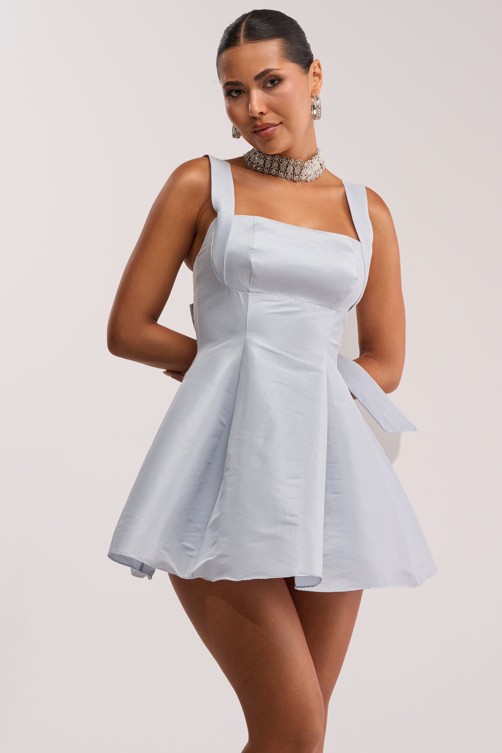 A woman models the BEST TIME OF THE YEAR SATIN MINI DRESS IN BABY BLUE, featuring a feminine silhouette. She stands with her hands behind her back, styled with a sleek hairstyle and statement choker necklace.