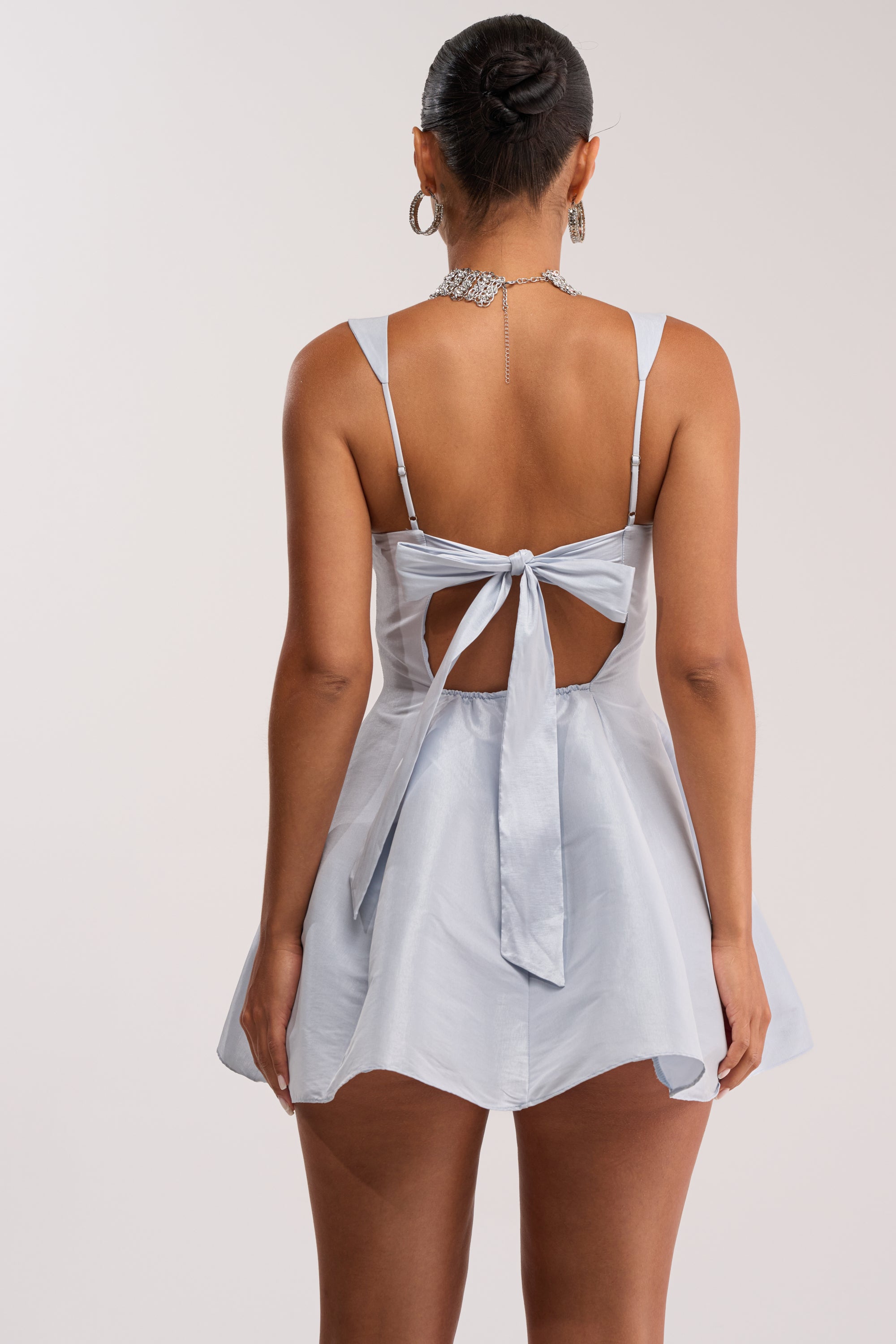 A woman wears the BEST TIME OF THE YEAR SATIN MINI DRESS IN BABY BLUE, featuring a flared skirt, open back with a large bow, her hair in a bun, and accessorized with hoop earrings and a sparkly necklace for a feminine look.
