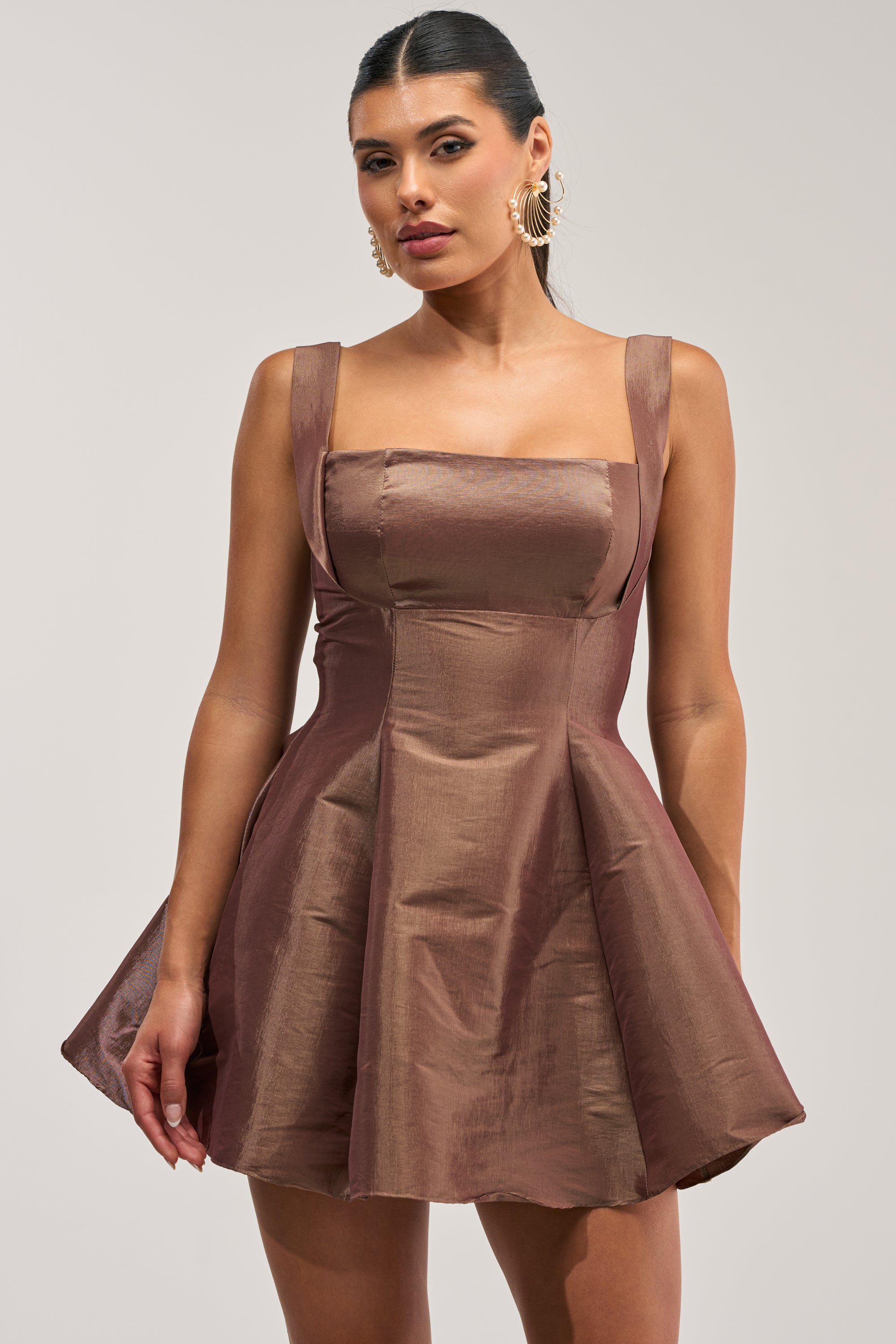 A woman wears the BEST TIME OF THE YEAR SATIN MINI DRESS IN MOCHA, a brown fit-and-flare satin mini dress featuring a square neckline and wide straps, styled as the perfect homecoming dress. She pairs it with gold shell pearl earrings against a light gray background.