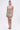 A woman models the COME MY WAY RUFFLE MINI DRESS, a strapless beige mini with layered ruffles and an uneven hem. Her hair is tied up and she pairs the dress with strappy sandals, standing against a plain white background.