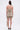 A person with long dark hair in a half-up style wears the COME MY WAY RUFFLE MINI DRESS—a strapless beige mini with ruched, layered ruffles and an asymmetrical train—facing away, showing an adhesive medical patch on their upper left arm.