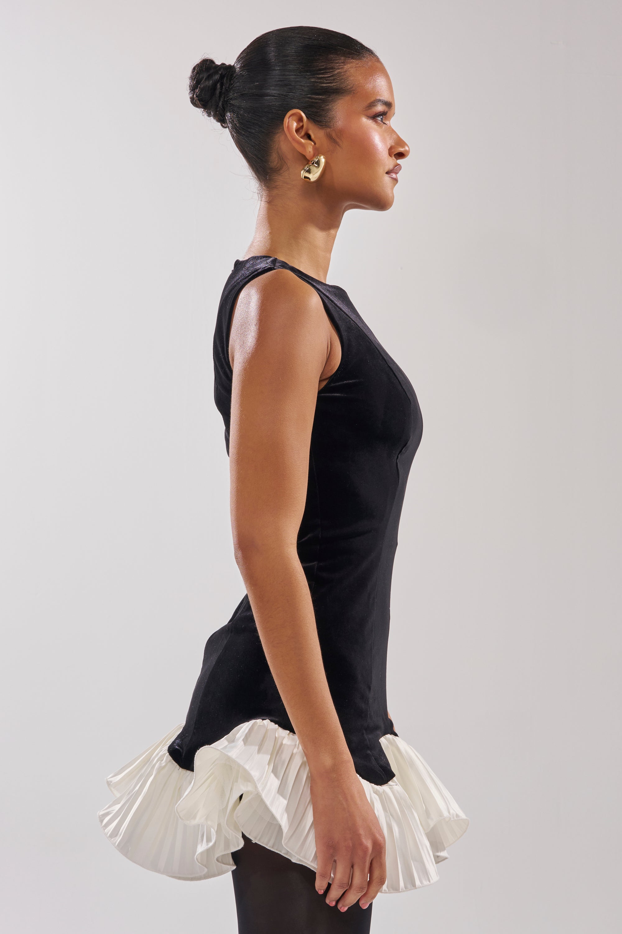 A woman stands in profile against a plain background, wearing the BLACK SWAN VELVET MINI DRESS—a sleeveless, high-neck style with a white ruffled hem—paired with black tights, gold hoop earrings, and her hair in a neat bun.