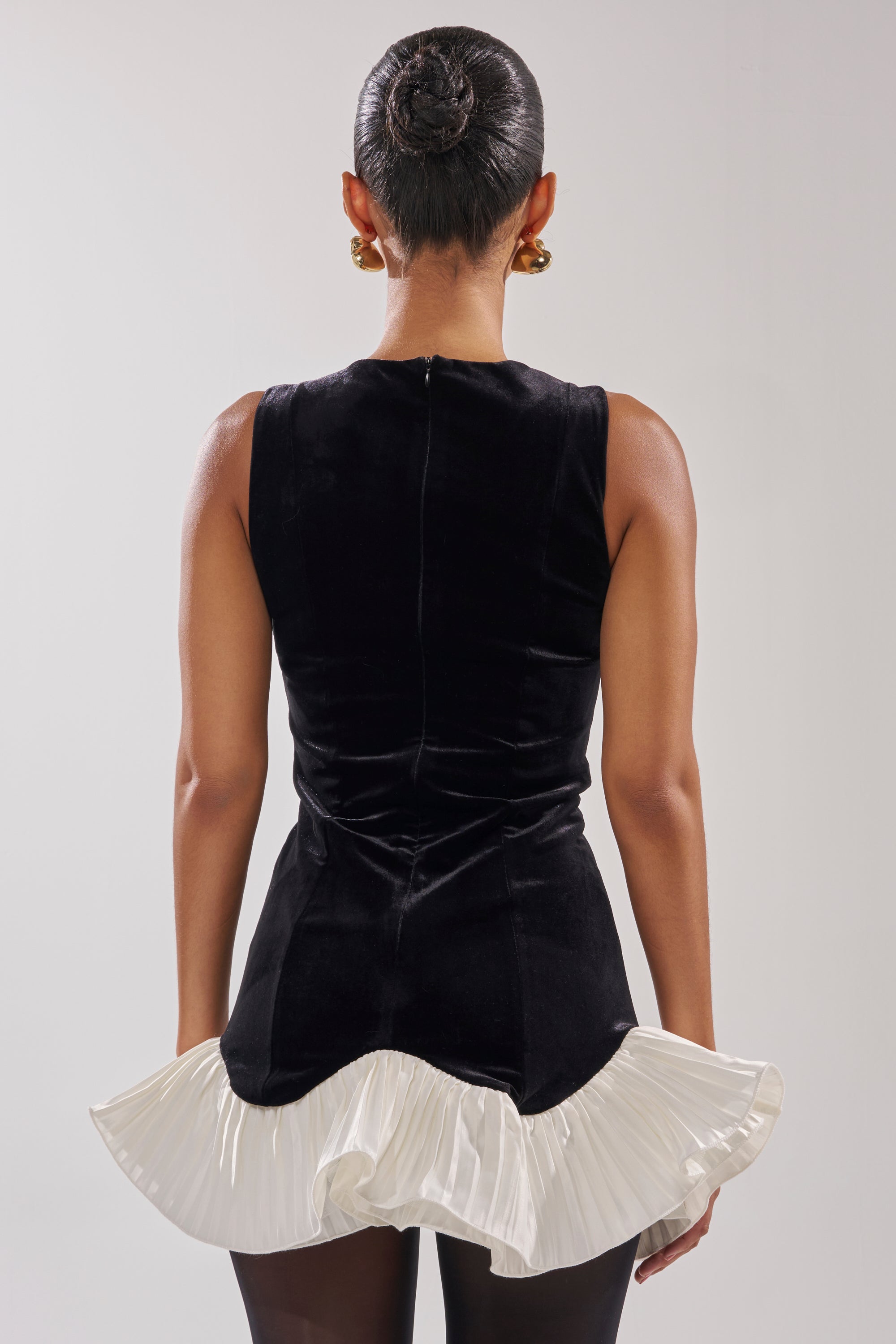 A woman with slicked-back hair in a bun models the BLACK SWAN VELVET MINI DRESS—sleeveless, black velvet with a white pleated ruffle hem—paired with gold earrings and black tights, shown from the back against a plain background.