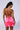 A woman with dark, wavy hair in a ponytail is showing off the vibrant neon pink IM A VIBE SLINKY MINI DRESS. She is facing away from the camera, highlighting the backless design with criss-crossed straps and tie detail.