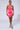 A woman stands against a plain background wearing the IM A VIBE SLINKY MINI DRESS in shiny, neon pink. She has her hair pulled back and is accessorized with a silver necklace and sparkling high heels.