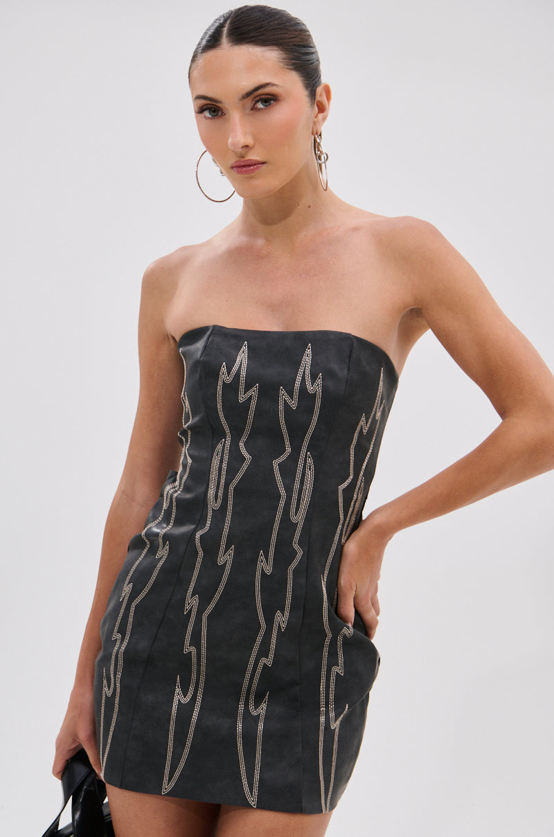 A woman models the REAL LIFE BOOGIE PU MINI DRESS, a strapless dress with bold wavy patterns. She pairs it with hoop earrings, slicked-back hair, and a small black bag for a chic, modern vibe against a plain background.