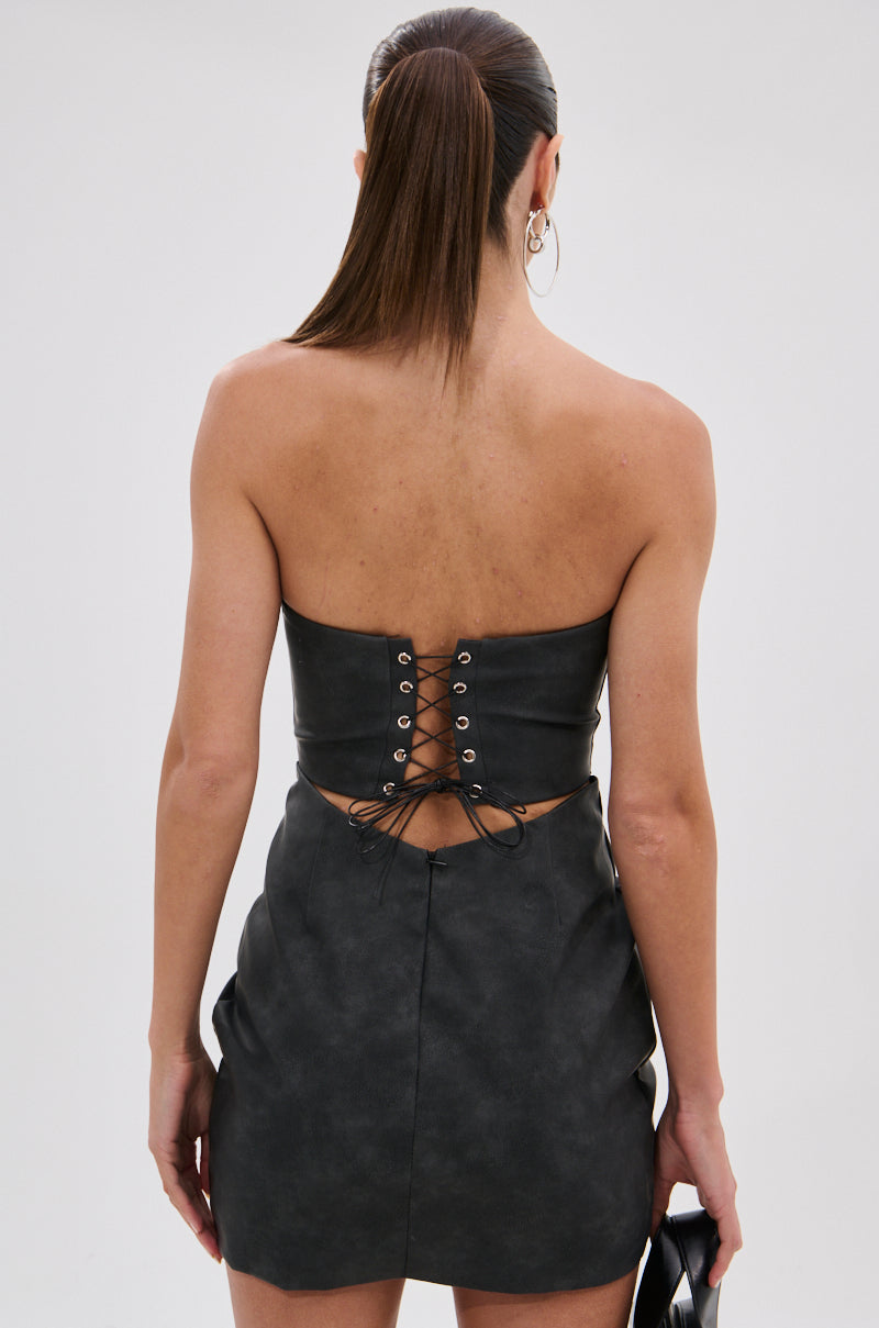 A woman with straight brown hair in a ponytail wears the REAL LIFE BOOGIE PU MINI DRESS, a strapless faux leather bodycon mini with lace-up back detail. She is facing away, holding a small black handbag.