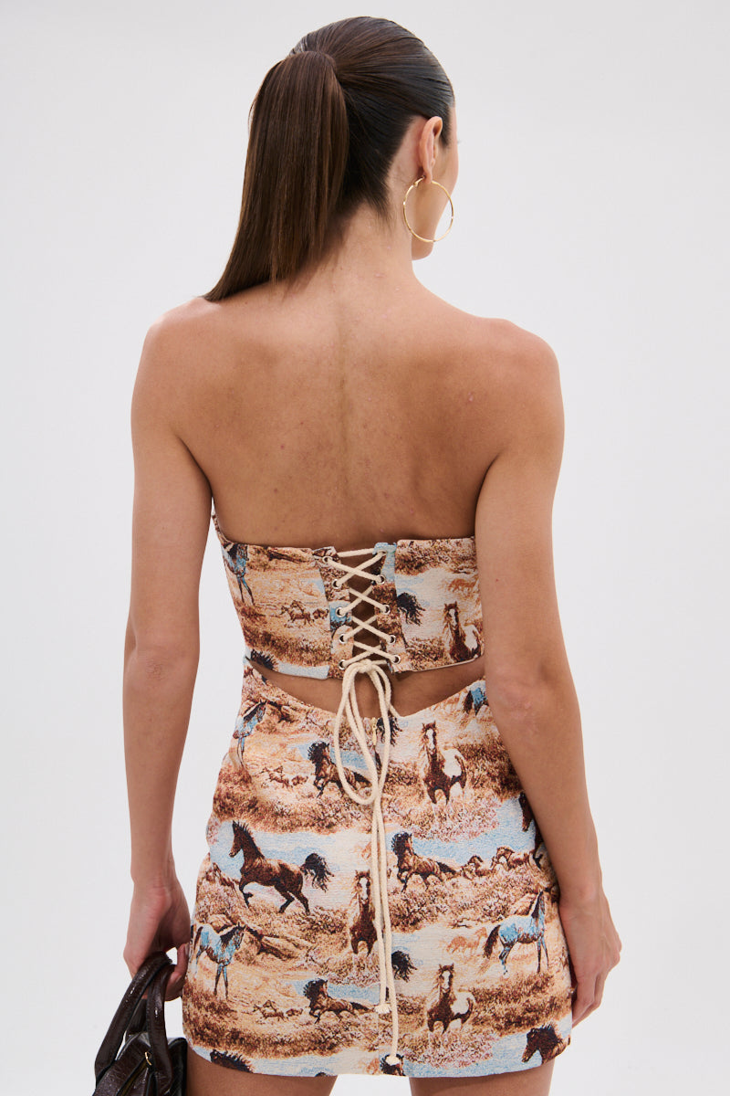 A woman with long, straight brown hair in a ponytail wears the AINT NO HOLD EM PRINTED MINI DRESS, featuring a Western horse print and lace-up back. Holding a dark bag, she faces away to reveal the dress’s striking back and bodycon fit. 5
