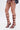 A person in a colorful pleated skirt wears AZALEA WANG DAGON LIQUID LUXE CAGED SANDALS—burgundy faux patent leather, strappy, with flared stiletto heels and open square toes—while standing against a plain white background.