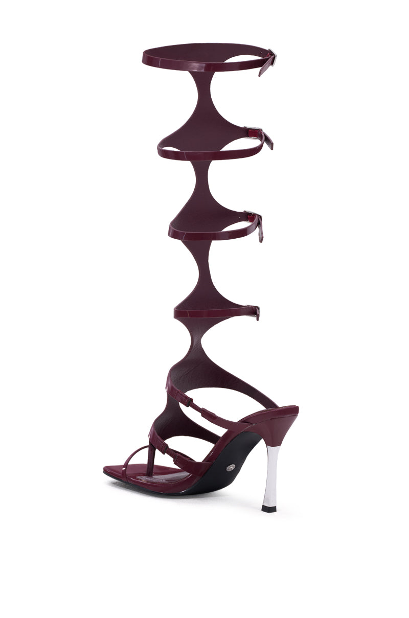 The AZALEA WANG DAGON LIQUID LUXE CAGED SANDAL features dark burgundy faux patent leather, an open square toe, sculptural strappy design with circular cutouts, multiple ankle straps, a wrap-up leg style, and a contrasting white heel.