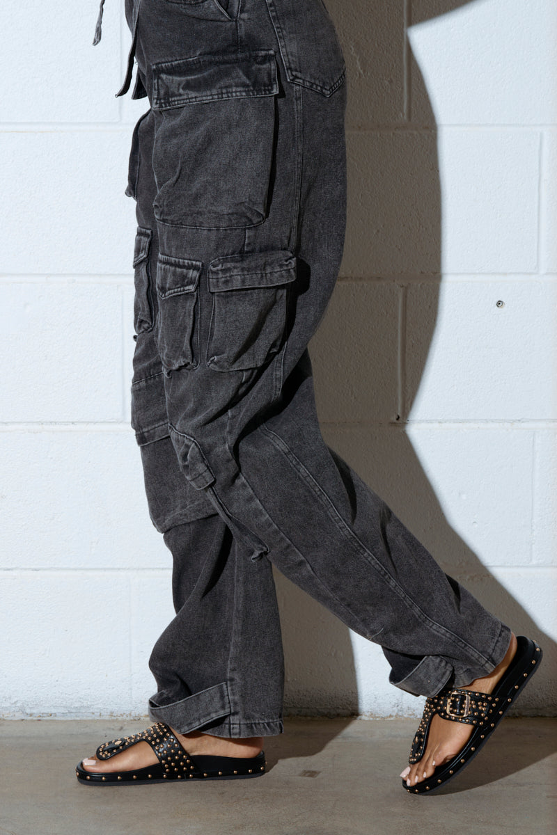 A person in dark gray cargo pants and AZALEA WANG DAISY CHAIN STUDDED BLACK SANDAL walks on a polished concrete floor. The white textured wall in the background highlights the movement-focused shot of their lower half.