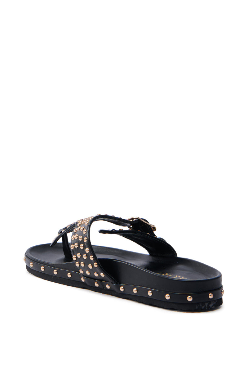 The AZALEA WANG DAISY CHAIN STUDDED BLACK SANDAL is a chic faux leather sandal with metallic gold studs on crisscross straps and along the sole edge, featuring a flat design. It’s stylishly shown from a slight rear angle against a white background.