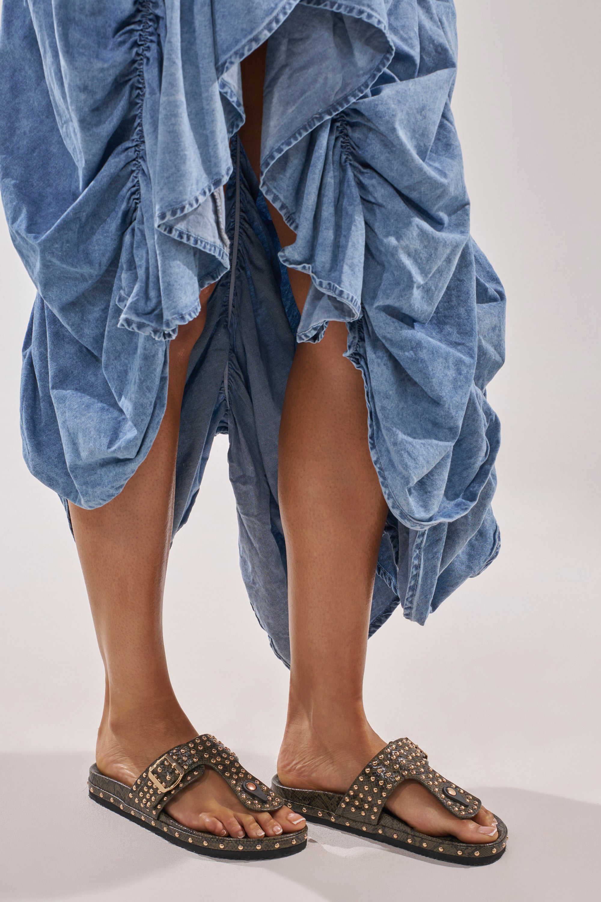 A person stands against a plain, light background wearing a blue, ruffled, knee-length skirt and AZALEA WANG DAISY CHAIN GREEN SNAKE STUDDED FLAT SANDALS. 1
