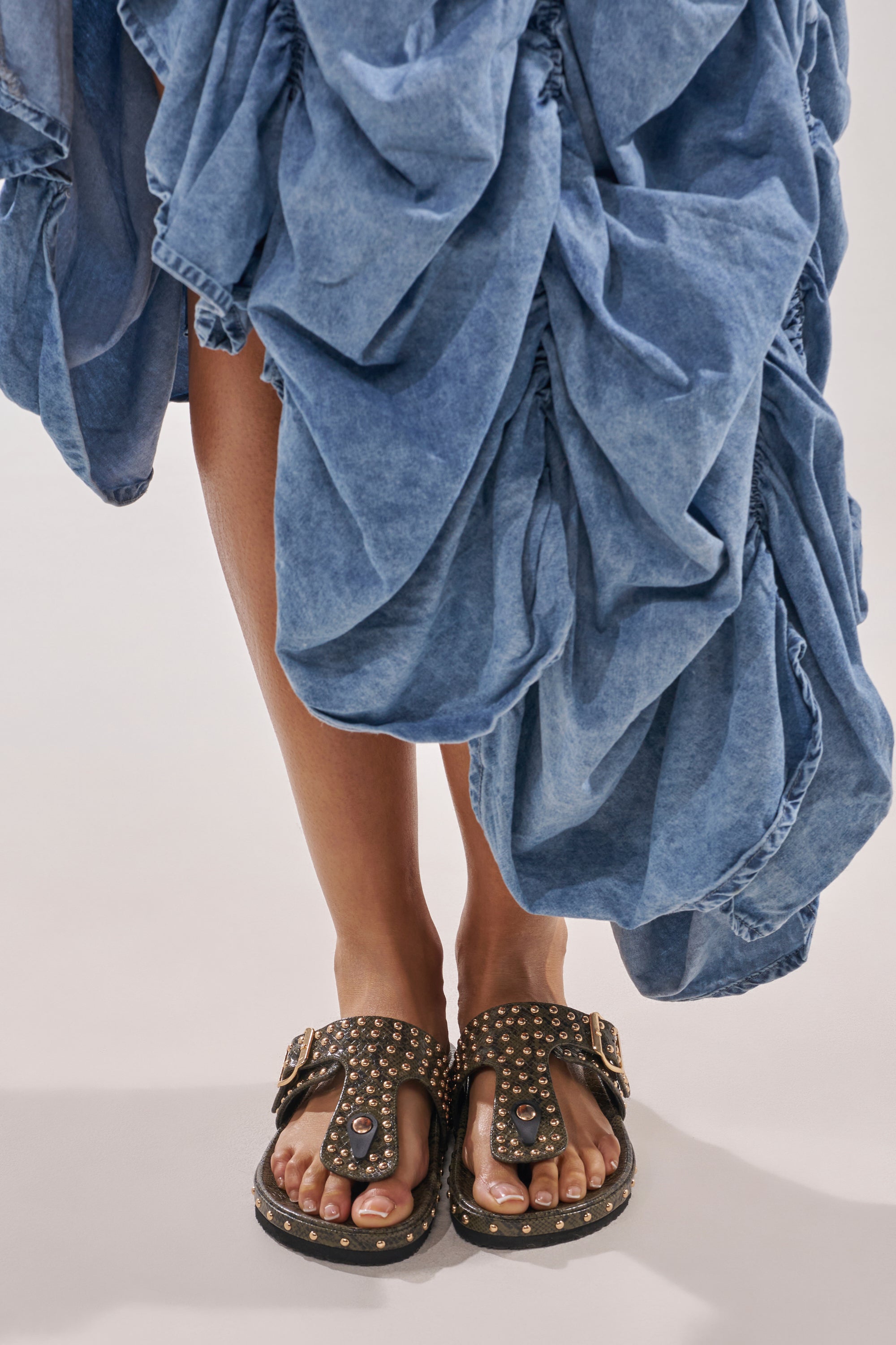 A person wears a blue, ruffled, denim-like skirt and stands with their feet visible in AZALEA WANG DAISY CHAIN GREEN SNAKE STUDDED FLAT SANDALS featuring gold studs and large buckles. 2