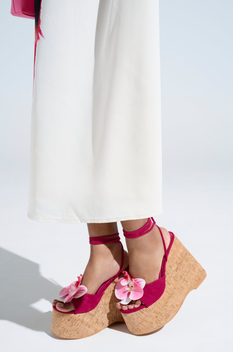 A person is wearing long white pants and the AZALEA WANG DALEYZA FUCHSIA FLORAL WEDGE SANDALS, which feature pink platform sandals with a fuchsia floral design, cork wedge chunky heels, suede textile upper, and delicate ankle straps. The background is plain and well-lit.