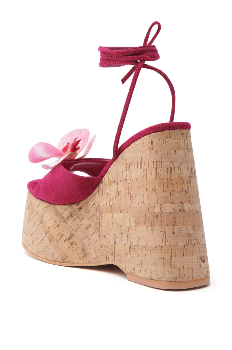 The AZALEA WANG DALEYZA FUCHSIA FLORAL WEDGE SANDAL showcases a side view of its fuchsia suede textile upper, highlighted by a large pink flower embellishment, elegant wrap-around ankle strap, and chunky cork heel.