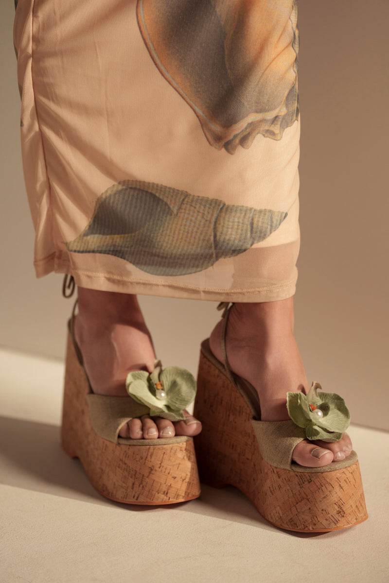 A person stands on a light surface wearing a peach-printed skirt and AZALEA WANG DALEYZA GREEN FLORAL WEDGE SANDALS featuring a canvas upper and cork wedge heel.