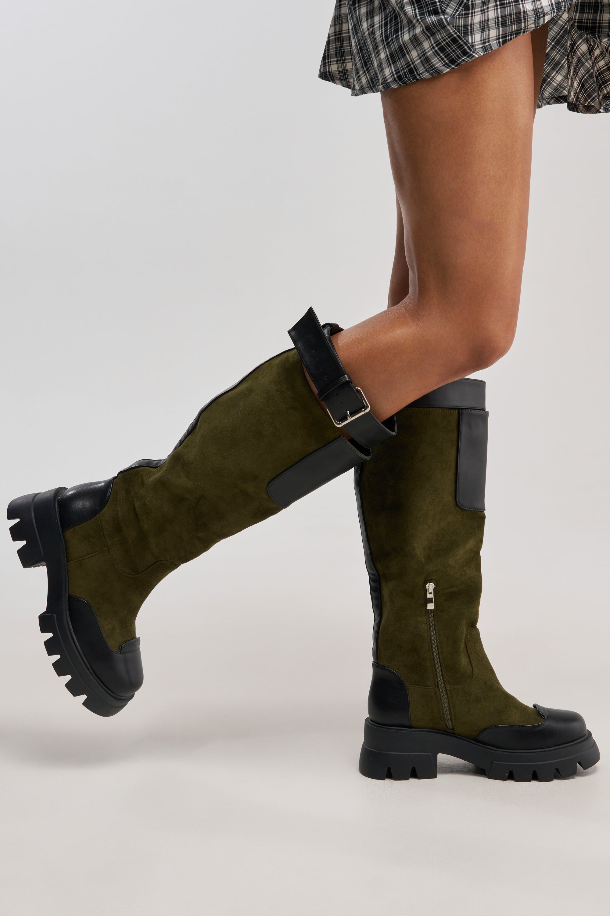 A person wears the AZALEA WANG DALILA OLIVE FAUX SUEDE FLAT BOOT—olive green knee-high faux suede boots with chunky black soles, black toe and heel accents, a side zipper, and top buckle—paired with a plaid skirt.