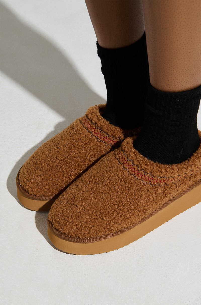 A person wears cozy AZALEA WANG DAMARI CHESTNUT FLAT slippers, featuring a rounded design with a small decorative stripe, over black socks on a light floor.