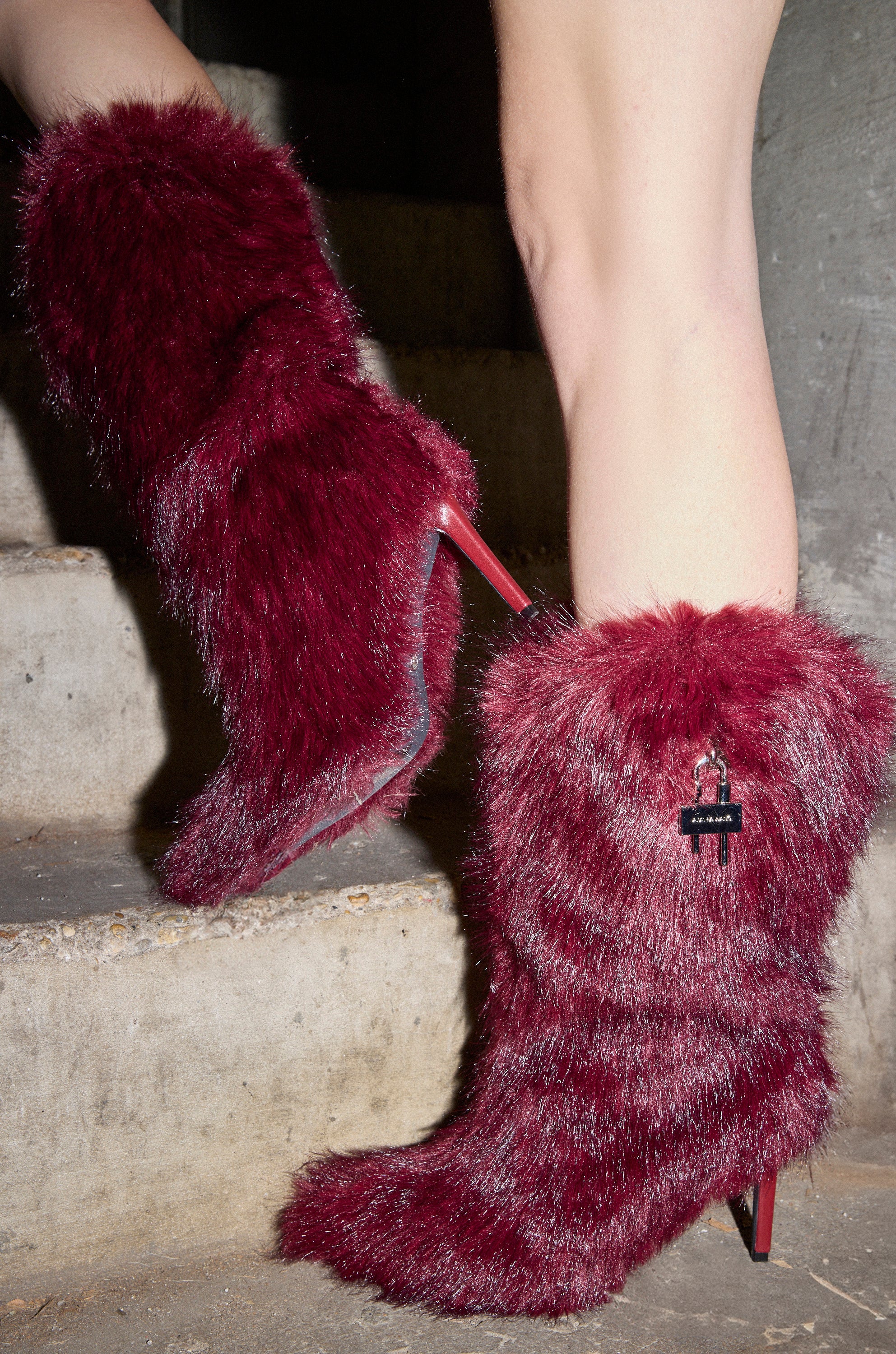 Someone wearing the AZALEA WANG DAMME RED FUR BOOTIE WITH LOCK DETAIL stands on concrete steps. One tall, faux fur stiletto boot in dark red is lifted, showing a small metallic lock decoration on the side.