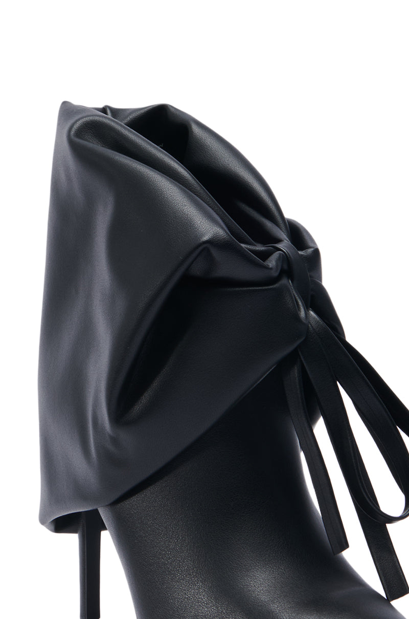 Close-up of the AZALEA WANG DARALEY STILETTO BOOTIE, a black high-heeled shoe adorned with an elaborate large bow on the side. It features sleek and shiny faux leather material and a pointed toe, offering a sophisticated and elegant appearance.