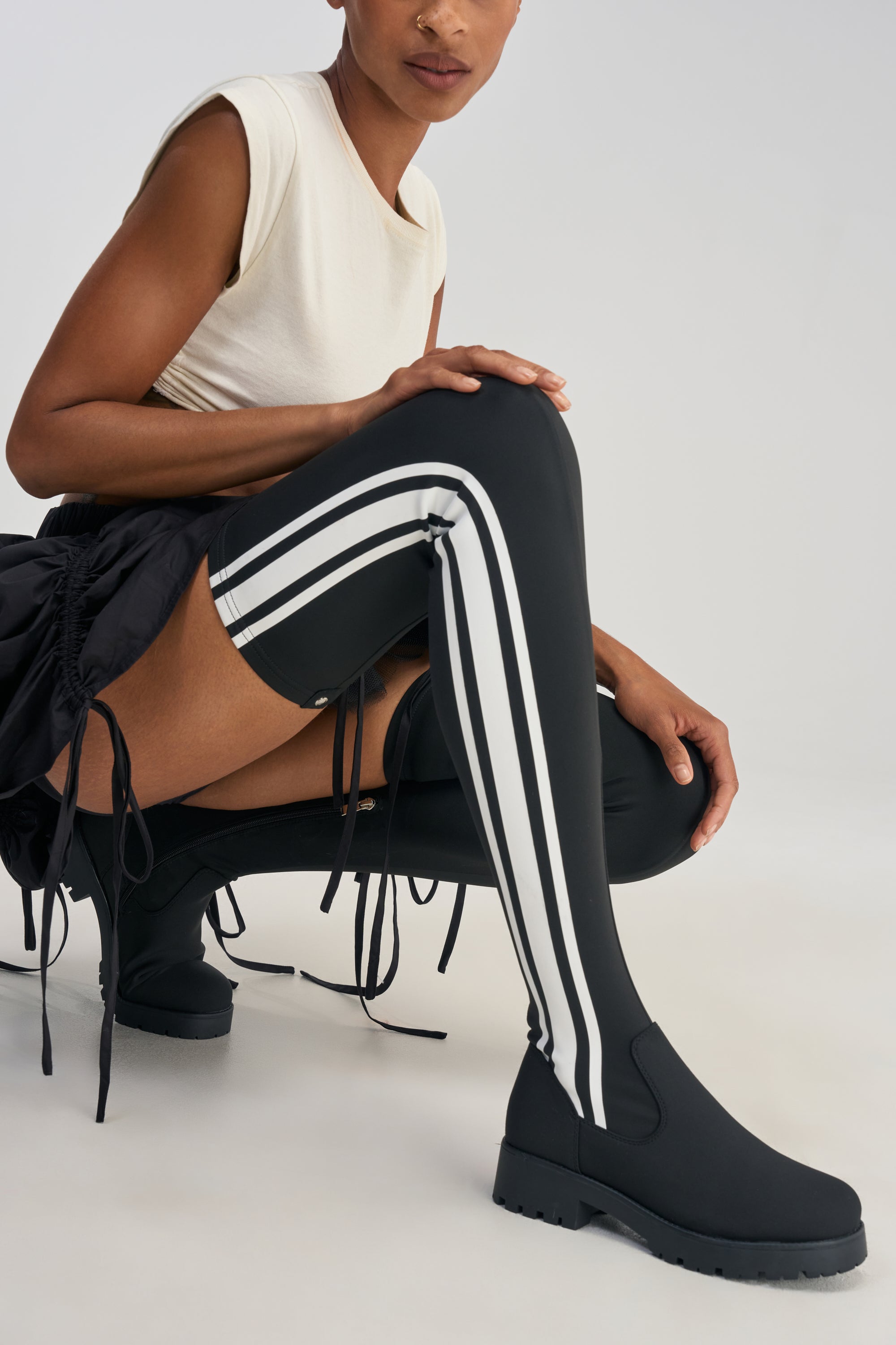 Wearing the AKIRA GOLD DARBY BLACK FLAT STRETCH SPORTY BOOT, a person kneels against a plain background, styled with black thigh-high tights featuring white stripes, a white sleeveless top, and a black skirt.