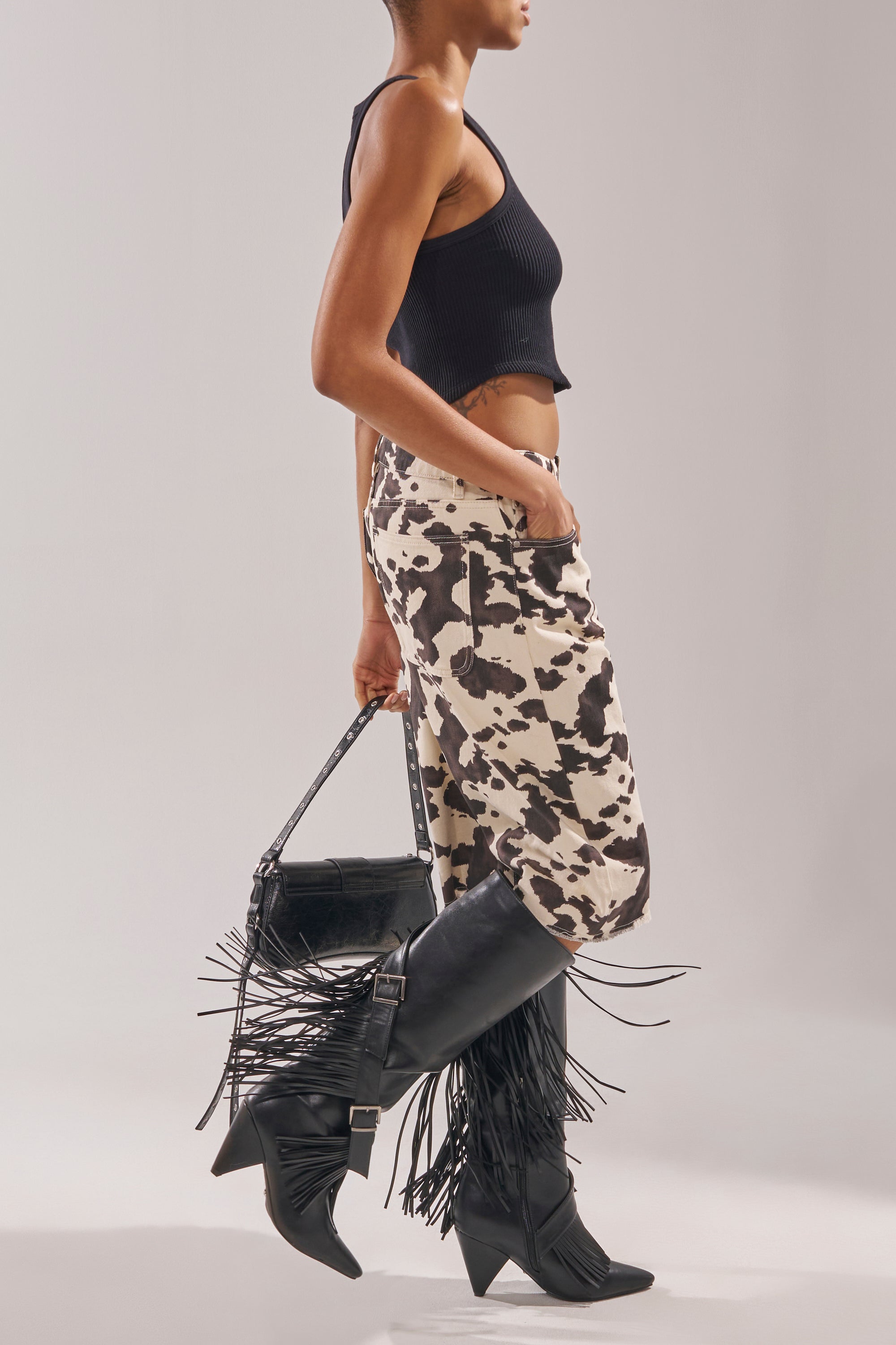 A person stands against a plain background, wearing a black sleeveless crop top, black and white cow print pants, and carrying a black fringe handbag while sporting the AZALEA WANG DARELLE BLACK FRINGE BOOT. 2
