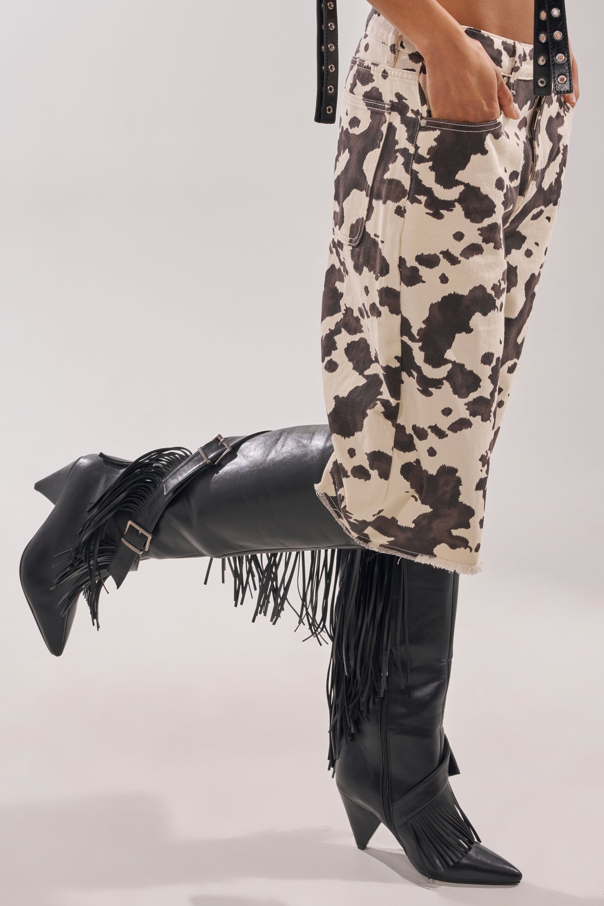A person wears the AZALEA WANG DARELLE BLACK FRINGE BOOT and cream shorts with a brown cow print, standing with one leg bent and a hand in their pocket. 3
