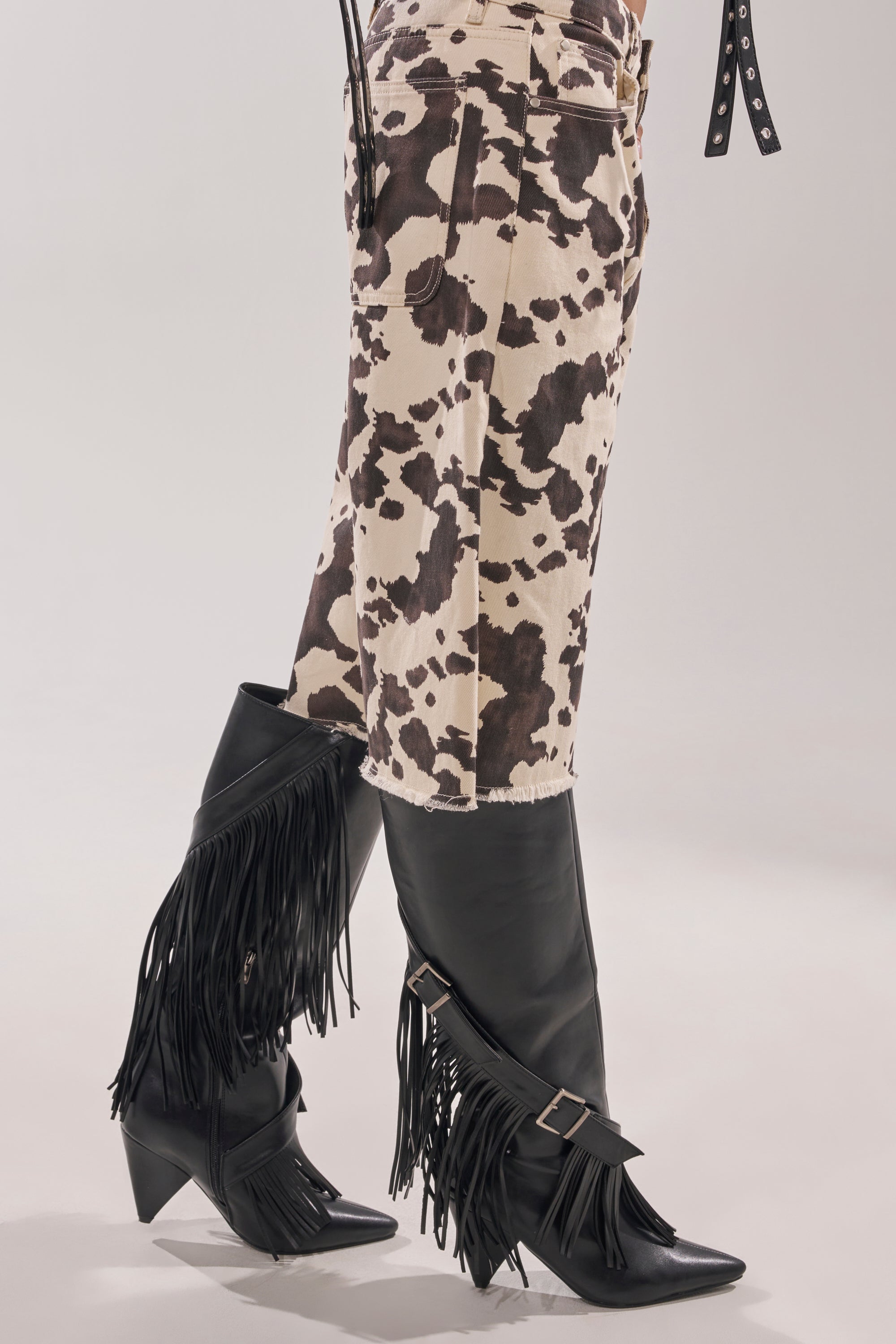 A person wears cow-print pants with AZALEA WANG DARELLE BLACK FRINGE BOOTS—knee-high, pointed-toe western boots with buckles—shown waist down against a plain light background. 8